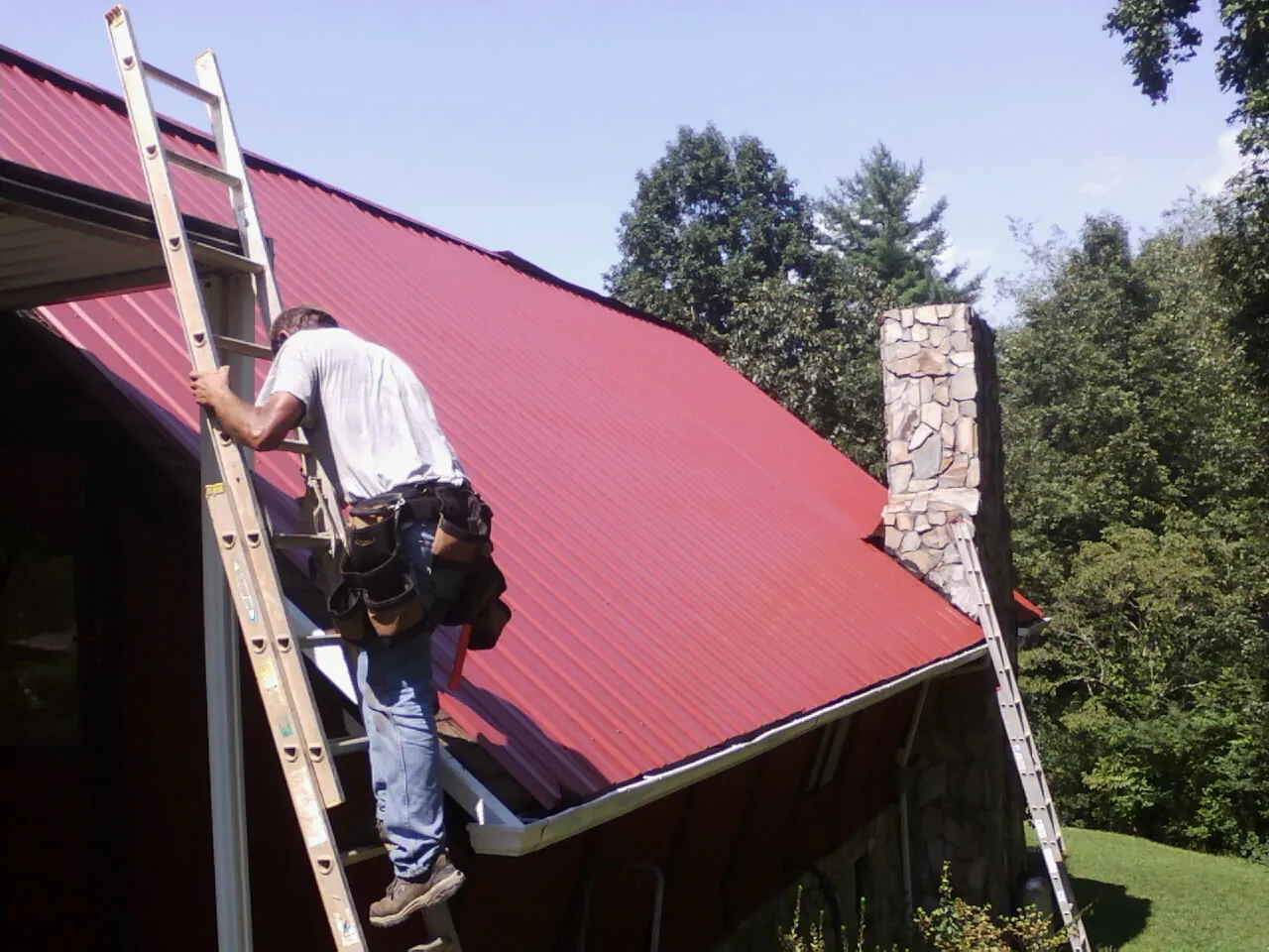 Roofer on a ladder beside a red metal roof and stone chimney — Soffit & Fascia Repair by Roofing Brothers in Bowdon